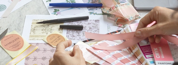 Close-up van handen die werken aan Mathilde M ontwerpen, kleurstalen en parfumlabels in een creatief atelier